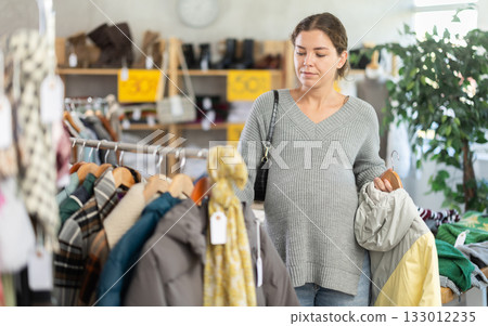Pregnant girl is busy looking at clothes items in store, she chooses coat or jacket 133012235