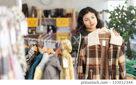 Stylish latina young woman standing in clothing shop and looking new coat or jacket made of high-quality material 133012344