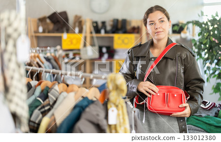 Fashionable european female customer standing near shelves with clothes in store and choosing handbag Fashionable european female customer standing near shelves with clothes in store and choosing handbag 133012380