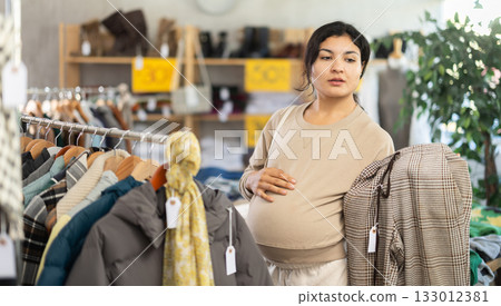 Pregnant mulatto young female client choosing trousers in clothing mall Pregnant mulatto young female client choosing trousers in clothing mall 133012381