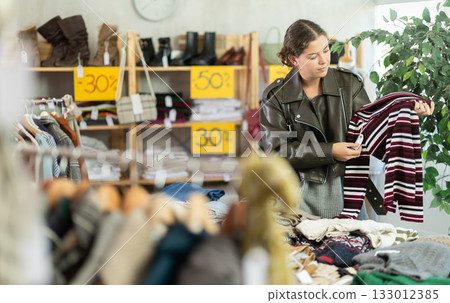 European girl is busy looking at clothes items in store, chooses sweater 133012385