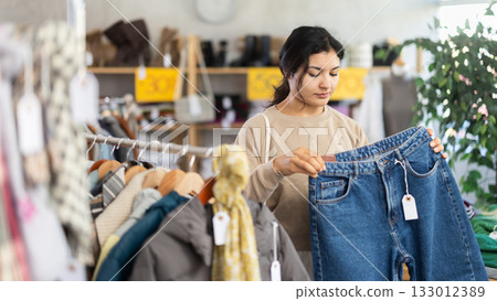 Happy latina female client carefully choosing stylish jeans trousers in clothing store. Shopping and sales concept Happy latina female client carefully choosing stylish jeans trousers in clothing store. Shopping and sales concept 133012389