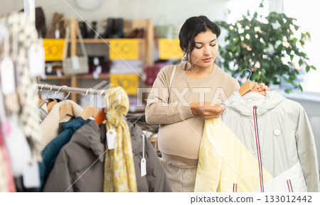 Pregnant Latin girl is busy looking at clothes items in store, she chooses winter coat or jacket 133012442
