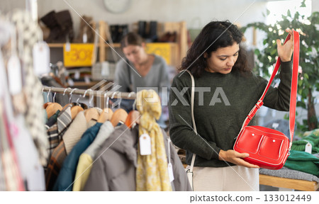 Happy latina woman standing in interior of clothing boutique and choosing fashionable handbag. Winter collection sale Happy latina woman standing in interior of clothing boutique and choosing fashionable handbag. Winter collection sale 133012449