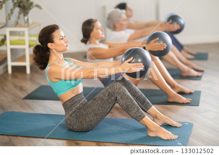 Women maintaining active lifestyle exercising with ball sitting on yoga mat during group workout in fitness 133012502