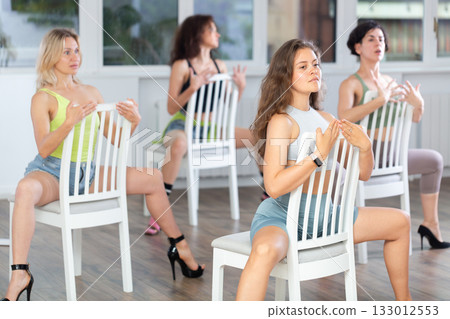 Sexy fit caucasian smiling woman demonstrating series of show-stopping stiletto dance moves sitting on chair, training with group of young women in dance hall 133012553