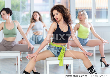 Sexy fit caucasian smiling woman demonstrating series of show-stopping stiletto dance moves sitting on chair, training with group of young women in dance hall Sexy fit caucasian smiling woman demonstrating series of show-stopping stiletto dance moves sitting on chair, training with group of young women in dance hall 133012566