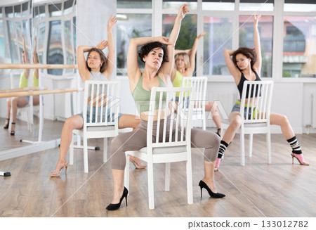 Beautiful energetic female trainer conducting classes in dance studio shows group correct performance of strip plastic exercise sitting on chair in studio 133012782