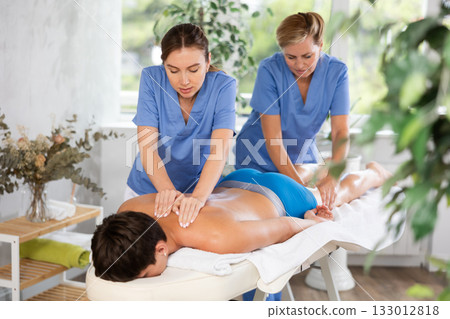 Two female masseuses perform stroking and rubbing of muscles of back and shoulders to guy client Two female masseuses perform stroking and rubbing of muscles of back and shoulders to guy client 133012818