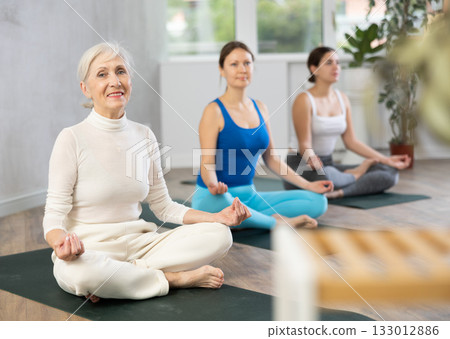 Women of different ages in sportswear relaxing in Lotus Posture Padmasana while practicing yoga class on retreat 133012886