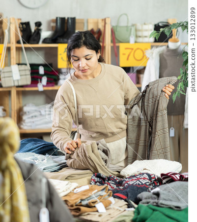 Latin girl is busy looking at clothes items in store, she chooses trousers 133012899