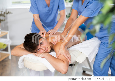 Two unrecognizable female masseuses perform stroking and rubbing of muscles of back to guy client 133012948
