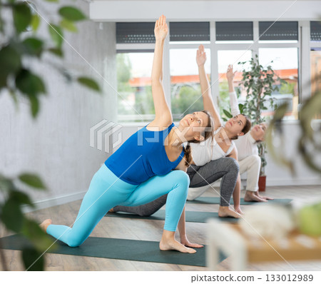 Women of different ages stretching in Extended Side Angle Pose or Utthita Parsvakonasana during yoga class in gym studio 133012989