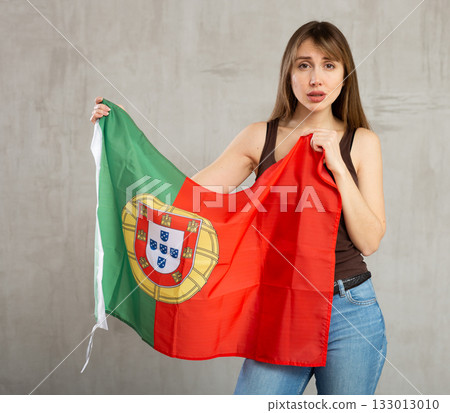 Sad young woman holding flag of Portugal against unicoloured background 133013010
