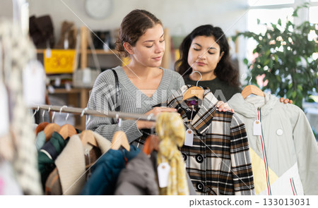Two happy young women choosing coat or jacket for cold season in clothing shop with large assortment Two happy young women choosing coat or jacket for cold season in clothing shop with large assortment 133013031