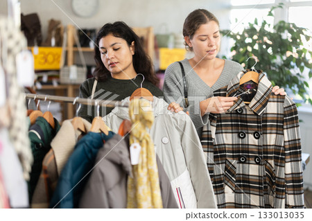 Two happy young women choosing coat or jacket for cold season in clothing shop with large assortment Two happy young women choosing coat or jacket for cold season in clothing shop with large assortment 133013035