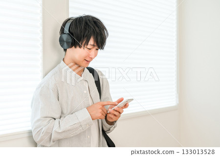 A young Asian man listening to music with headphones while operating his smartphone (music, subscription, app) 133013528