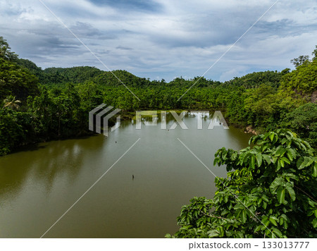 A calm freshwater lake is surrounded by dense tropical forest and hills under a cloudy sky. Siargao, Philippines. 133013777