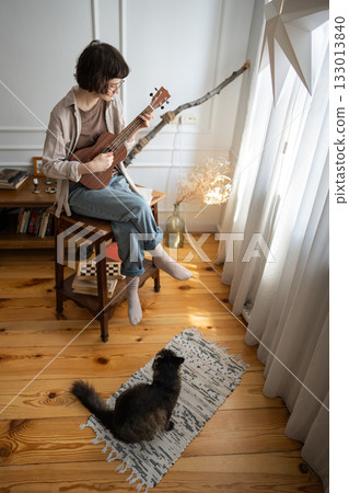 Immersed young woman musician practicing ukulele at home, observed by attentive household cat 133013840