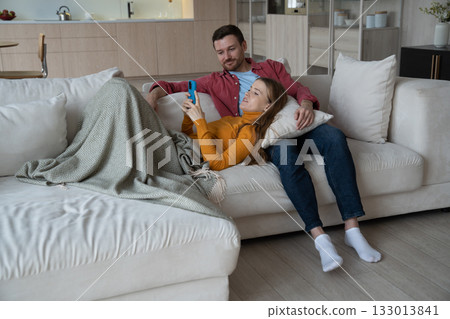 Cheerful woman lying on man knees, holding smartphone, smiling while having videocall with relatives 133013841