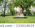 Asian woman meditating in a peaceful park setting, sitting cross-legged on green grass 133014639