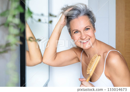 European beautiful and smiling middle-aged woman combing her gray hair near the mirror European beautiful and smiling middle-aged woman combing her gray hair near the mirror 133014673