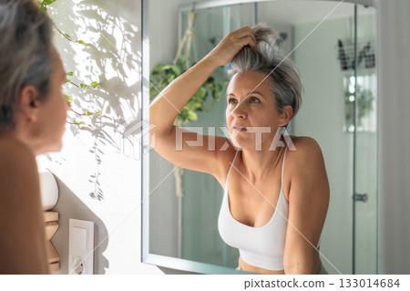 European beautiful middle-aged woman carefully examines her gray hair and wrinkles in the mirror. 133014684