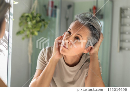 European white-skinned middle-aged woman looks anxiously in the bathroom mirror. Gray hair, wrinkles 133014696