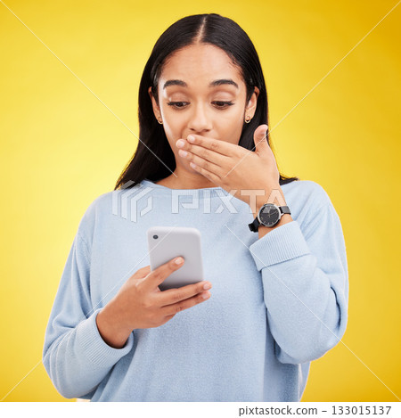 Shock, surprise and woman in studio with phone and hand on mouth isolated on yellow background. Social media, fake news or exciting online promotion, hispanic girl reading notification on smartphone. 133015137