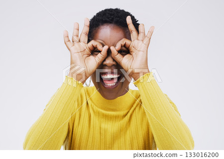 Portrait, funny face and finger glasses with a black woman laughing in studio on a white background for humor. Comic, comedy and hands on eyes with a playful young female person joking or having fun 133016200