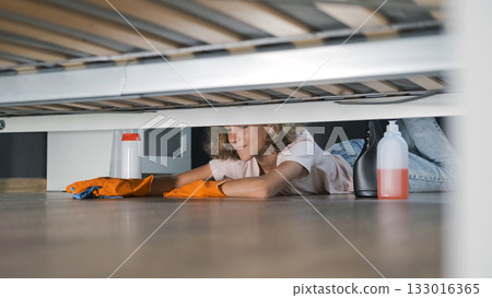 Teen girl lies on the floor cleaning under the bed in her room using cleaning supplies Teen girl lies on the floor cleaning under the bed in her room using cleaning supplies 133016365