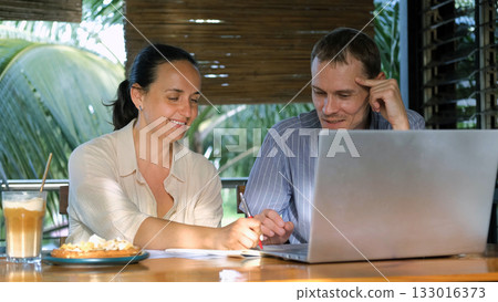 A man and woman discuss a new project working on a coffee shop terrace using a laptop and taking notes with drinks and snacks  133016373