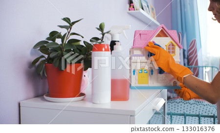 Teen girl wearing gloves wipes colorful toy house in a cozy space while doing chores Teen girl wearing gloves wipes colorful toy house in a cozy space while doing chores 133016376