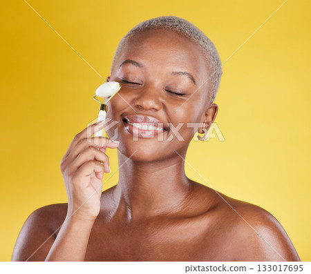 Face, skincare smile and black woman with roller in studio isolated on a yellow background. Beauty dermatology, massage and African female model with eyes closed and stone for facial skin treatment. 133017695