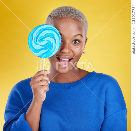 Excited, portrait and black woman with a lollipop in studio for dessert, sweet treat or craving. Happy, surprise and face of African female model posing with blue candy isolated by yellow background. Excited, portrait and black woman with a lollipop in studio for dessert, sweet treat or craving. Happy, surprise and face of African female model posing with blue candy isolated by yellow background. 133017784