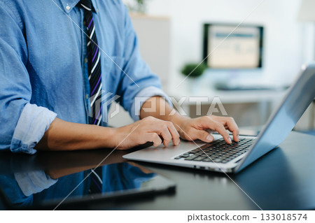 Young man typing on laptop and smartphone at modern office desk with coffee and tablet nearby. 133018574