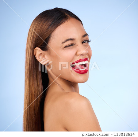 Beauty, woman and wink for face makeup and skincare cosmetics in studio. Female model person with tongue on teeth and red lips on a blue background for self care, facial glow and healthy skin shine Beauty, woman and wink for face makeup and skincare cosmetics in studio. Female model person with tongue on teeth and red lips on a blue background for self care, facial glow and healthy skin shine 133018634