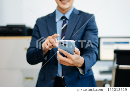 Smiling businessman in suit holds smartphone while gesturing to interact, symbolizing fintech, mobile ap 133018819