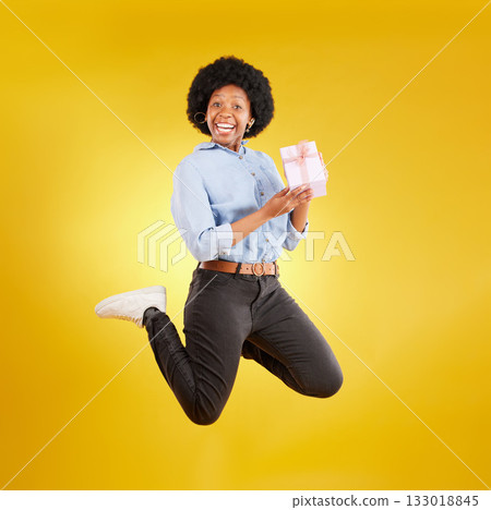 Jump, excited and black woman with gift in air on yellow background with energy, happy and smile in studio. Birthday mockup, celebrate and isolated girl jumping with present, box and surprise package 133018845