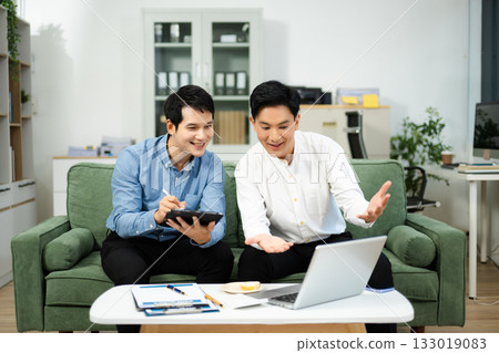 Happy Asian business partners collaborating on startup ideas using tablet and laptop in modern office. 133019083
