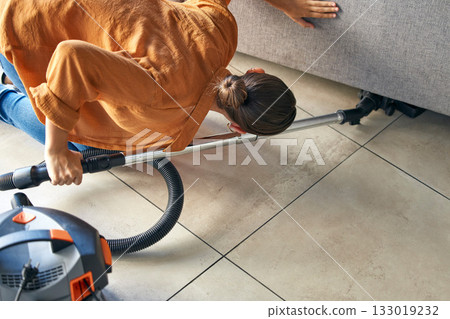 Woman vacuum cleaning under sofa in living room for housekeeping, hygiene maintenance or maid service. Cleaner, electric appliance and flooring, furniture dust and ground tiles and in lounge at home Woman vacuum cleaning under sofa in living room for housekeeping, hygiene maintenance or maid service. Cleaner, electric appliance and flooring, furniture dust and ground tiles and in lounge at home 133019232