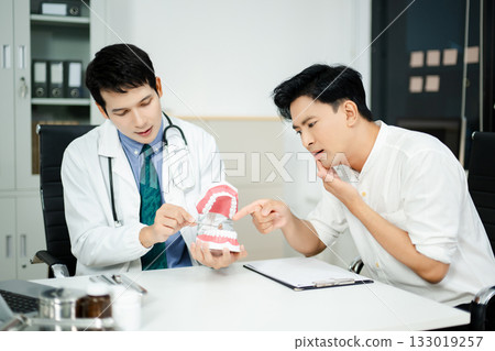 Dentist explaining dental health to male patient using model jaw. Oral care, tooth pain consultation, anatomy education Dentist explaining dental health to male patient using model jaw. Oral care, tooth pain consultation, anatomy education 133019257