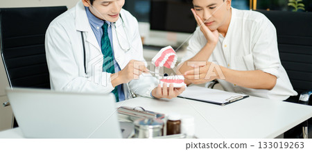 Dentist explaining dental care to male patient using a tooth model. Health consultation, oral hygiene, and professional support Dentist explaining dental care to male patient using a tooth model. Health consultation, oral hygiene, and professional support 133019263