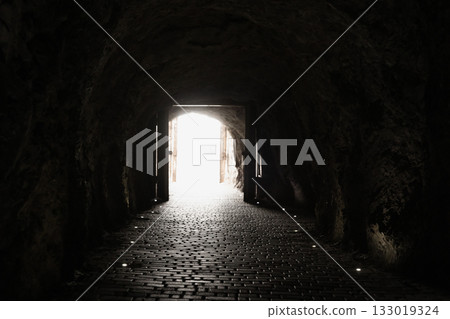 A dim underground tunnel with rough stone walls and a glowing exit at the end A dim underground tunnel with rough stone walls and a glowing exit at the end 133019324
