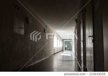 A quiet, dim corridor with closed doors on the right and natural light at the far end A quiet, dim corridor with closed doors on the right and natural light at the far end 133019326