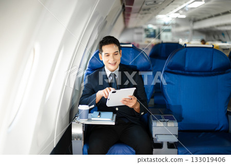 Confident businessman enjoying luxury air travel in first class, working on a tablet with a coffee, blending business, technology, and global connectivity. Confident businessman enjoying luxury air travel in first class, working on a tablet with a coffee, blending business, technology, and global connectivity. 133019406