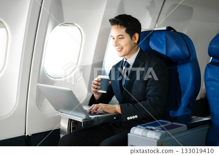 Confident businessman enjoying luxury air travel in first class, working on a tablet with a coffee, blending business, technology, and global connectivity. Confident businessman enjoying luxury air travel in first class, working on a tablet with a coffee, blending business, technology, and global connectivity. 133019410