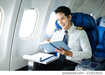 Asian Confident male pilot in uniform leaning at the passenger seat while standing inside of the airplane flight cockpit during takeoff and checking flight plan 133019523