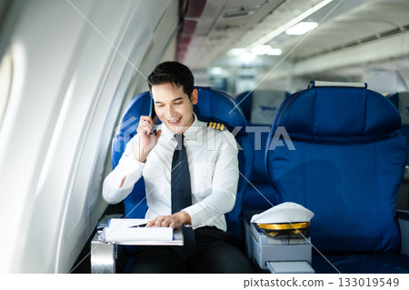 Confident airline pilot in uniform using wireless earbuds while reviewing documents, perfect for aviation, travel, and modern communication themes Confident airline pilot in uniform using wireless earbuds while reviewing documents, perfect for aviation, travel, and modern communication themes 133019549