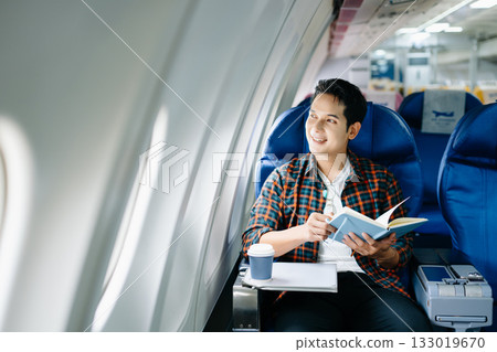 Happy young man traveling by plane. Business or leisure flight series showing phone talk, tech, reading, and stress. 133019670